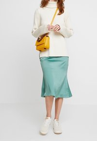 Pull à col roulé en crème à côtes, jupe midi satin teal, baskets blanches, sac à main jaune avec des détails floraux. Tenue simple et moderne.