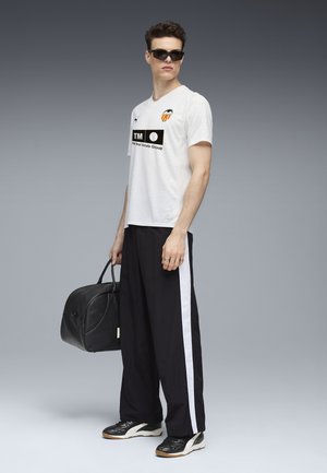 Witte shirt met korte mouwen en logo-accenten, zwarte trainingsbroek met een witte zijstreep, zwarte sneakers en een zwarte sporttas.