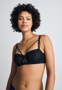 Black lace bra featuring embroidered cups, scalloped edges, and multiple strap designs with gold hardware accents.