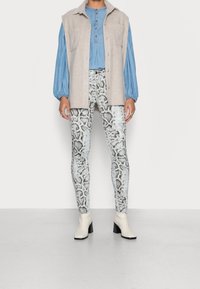 Beige, ärmlös väst över en blå blus, matchad med leggings i ormskinnstryck i vitt och grått. Modellen bär krämbestra ankle boots med blockklack.