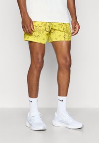 Bañador amarillo con un patrón paisley en marrón. Presenta un bolsillo lateral y un detalle del logo. Combinado con zapatillas blancas y calcetines.