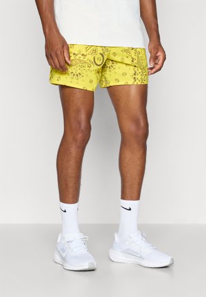 Gule svømmetights med et paisleymønster i brunt. Har en sidelomme og en logodetalj. Bæres med hvite joggesko og sokker.