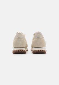 Beige suede athletic shoes with a low profile, textured material, flat rubber outsole, and subtle logo detailing on the heel.