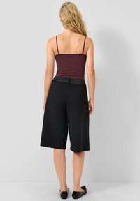 Kobieta z długimi, falującymi blond włosami, ubrana w bezrękawnik w kolorze bordowym, czarne szerokie szorty do kolan i czarne płaskie buty, stojąca pod jednolitą ścianą.