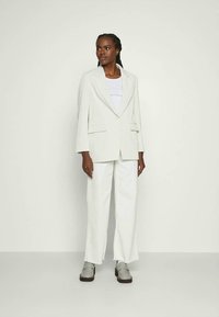 Lys grå blazer med en skreddersydd passform, kombinert med en hvit topp og vide, hvite bukser. Modellen har på seg grå loafers, som viser en ren, moderne stil.