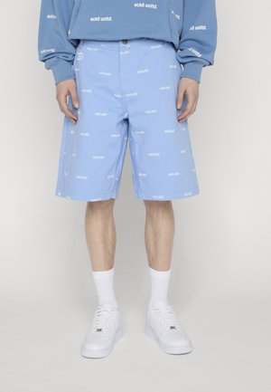 Pantalones cortos azules con logo blanco "eckō unltd." impresos, usados con sudadera a juego, calcetines blancos y zapatillas blancas en una persona de pie.
