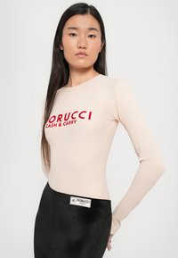 Dugih rukava, kožni bodi u nude boji s natpisom "FIORUCCI CASH & CARRY" u crvenom. Uparen s crnom suknjom visokog struka s brendiranim oznakom.