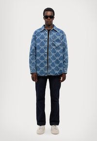 Blå denimskjorta med vågmönster, knappstängning och bröstficka, kombinerad med mörka jeans och ljusa sneakers. Solglasögon på.