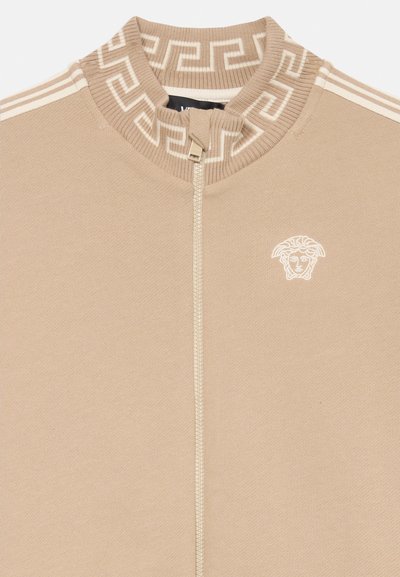 Beige Zip-Jacke mit Reißverschluss und geribkeltem Kragen, mit einem griechischen Schlüsselmotiv. Goldfarbene Hardware und ein weißes gesticktes Logo auf der Brust.