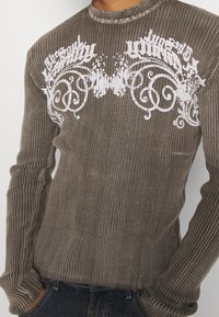 Brown ribbed long-sleeve sweater with white graphic print across the chest, featuring a round neck and fitted design.