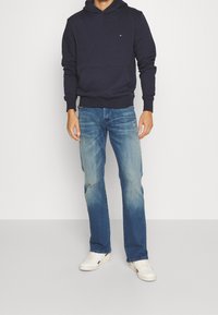 Námořnická mikina s kapucí s přední kapsou a žebrovanými manžetami, v kombinaci se světle modrými džíny s bledým efektem a bílými teniskami. Jednoduchý design, pohodlný střih.