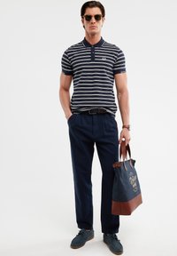 Polo cu dungi albastre și albe, cu guler, purtat cu pantaloni albaștri și loafers albaștri. Poartă o geantă de umăr bleumarin cu accente maro.