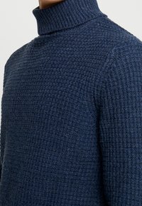 Mörkblå stickad polotröja med ett texturerat mönster och ribbad krage. Närbild som framhäver tygdetaljerna.