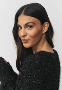 Black fuzzy sweater with metallic accents, loose fit, wide neckline, and long sleeves. Model's hair is dark and wavy, framing the face.