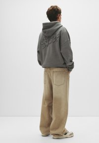 Grauer Kapuzenpullover mit einem Logo auf der Rückseite, kombiniert mit beigen weit geschnittenen Hosen. Das Outfit wird mit Sneakers mit schwarzen Akzenten abgerundet.