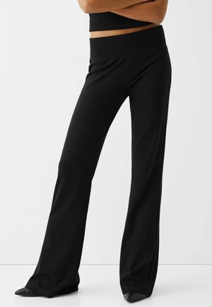 Mujer con pantalones negros de talle alto y pierna ancha y zapatos negros puntiagudos, de pie con los brazos cruzados frente a un fondo liso.