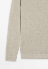 Beige strikket langærmet sweater med ribbet manchetter og kant, vist fra midt på armen til taljen.