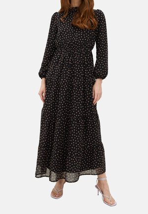 Kvinde iført en lang sort kjole med små blomsterprint, lange ærmer og flæsekant, kombineret med lyse lilla remsandaler med høje hæle.