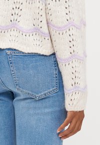 Sweater de malha em creme com padrão em ponto aberto, com detalhes em lilás com borda ondulada. Combinado com jeans de cintura alta azuis e um detalhe no bolso traseiro.
