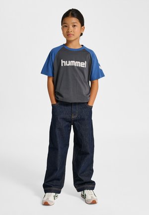 Szaro-niebieska koszulka typu raglan z białym logo "hummel", zestawiona z ciemnoniebieskimi dżinsami i białymi trampkami z zielonymi akcentami.