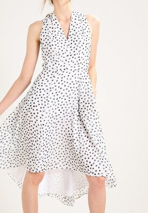 Mujer con vestido blanco sin mangas con lunares negros, bajo alto-bajo y escote en V, de pie con un brazo extendido.