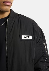 Giubbotto bomber nero con colletto a coste, chiusura con zip e una toppa etichettata "AVX". Presenta tasche laterali e una superficie liscia.