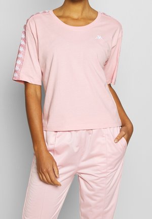 Kvinde iført en lyserød kortærmet skjorte med hvidt logo på brystet og mønstrede striber på ærmerne, kombineret med matchende lyserøde bukser.