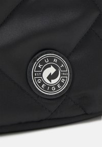 Mustalla päällysteellä varustettu tekstiili, jossa on pyöreä logopatchi, jossa on valkoinen nuolimuotoilu ja teksti "Kurt Geiger EST 1963" musta-valkoisena.