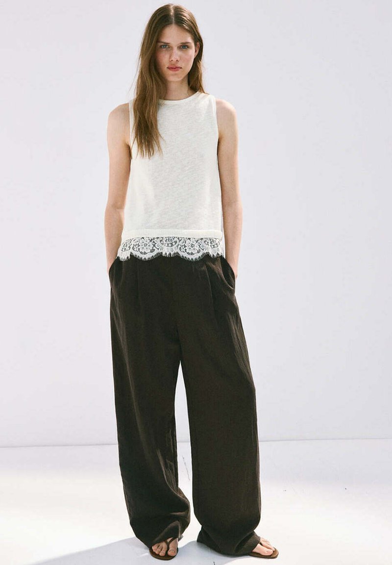 Jeune femme aux longs cheveux bruns portant un haut blanc sans manches avec un ourlet en dentelle et un pantalon noir ample, debout les mains dans les poches.