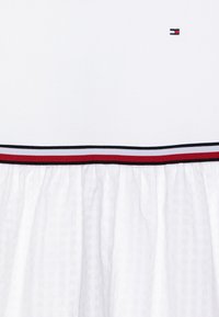 Weißes Kleid mit gerafftem Rock, mit einem horizontalen Kragen mit einem rot-weiß-schwarzen gestreiften Besatz und einem kleinen Logo auf dem Oberteil.