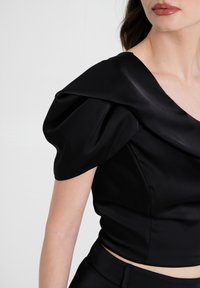 Top corto nero con maniche a sbuffo, realizzato in satin liscio. Presenta un profondo scollo a V e un corpo aderente e snello.