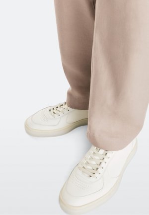 Voeten met witte sneakers en losse lichtbruine broek op een witte achtergrond, met zicht op schoen details en broekzomen.