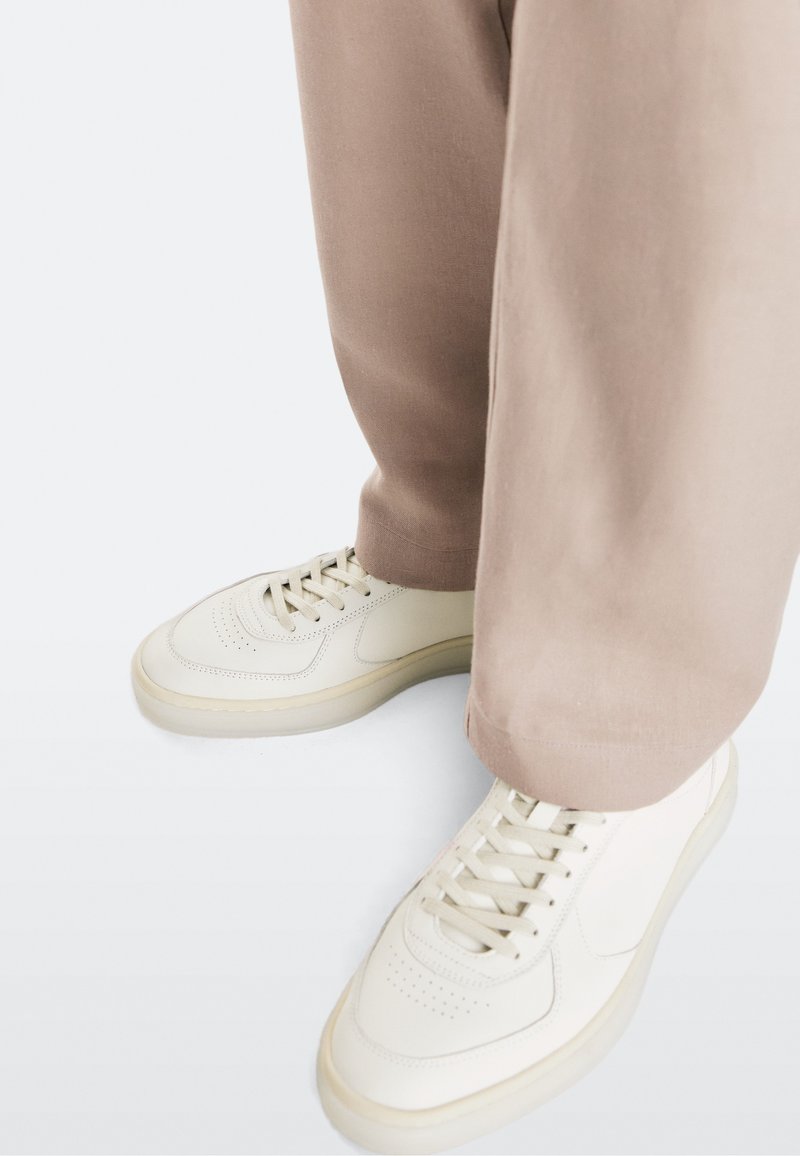 Feet wearing white sneakers and loose light brown pants on a white background, showing shoe details and pant hems.