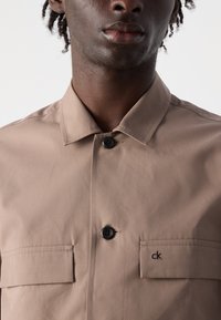 Tan button-up skjorte lavet af letvægtsstof, med klassisk krave, to frontlommer og broderet sort logo på venstre lomme.