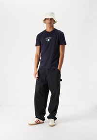 Marineblaues T-Shirt mit Grafikdruck, lässige schwarze Jeanshose, cremefarbene Sneaker mit roten Streifen und ein cremefarbener Bucket Hat.