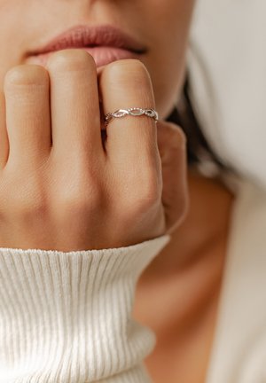 Anello d'argento con design intrecciato su un dito, caratterizzato da bande lisce e testurizzate. Il dito poggia su un indumento morbido e rigato.