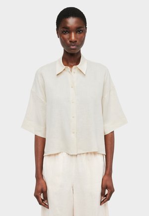 Frau trägt locker sitzendes cremefarbenes Kurzarmhemd mit Knöpfen und passende weit geschnittene Hosen, steht vor einem schlichten weißen Hintergrund.