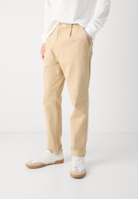 Beige színű laza nadrágot és fehér sportcipőt viselő személy, az egyik kezét zsebében tartva, sima háttér előtt áll.