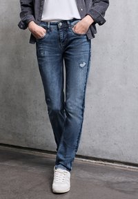 Siniset farkut slim fit -mallilla, joissa on kulutettu yksityiskohtia ja perinteinen viiden taskun malli. Yhdistetty valkoisiin lenkkitossuihin.