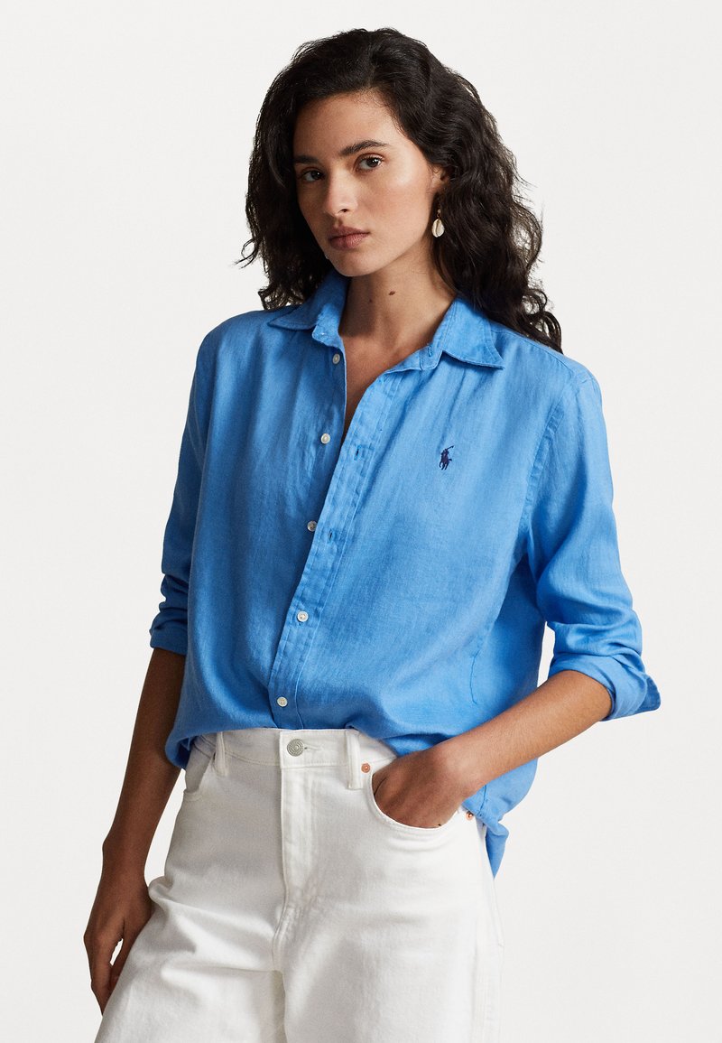 Light blue, linen button-up shirt with a collar, long sleeves rolled to the elbows, and subtle logo on the left chest. Paired with white pants.