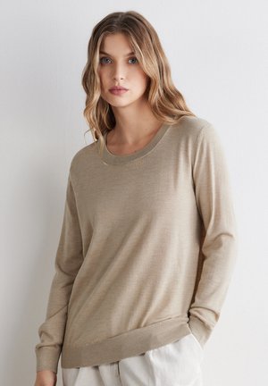 Kobieta o falowanych blond włosach, ubrana w beżowy sweter z długim rękawem i jasne spodnie, stojąca przy białej ścianie.