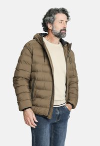 Chaqueta de plumas marrón con capucha, con costuras horizontales, bolsillos frontales con cremallera y detalles en negro. Usada sobre una camisa de color claro.