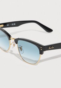 Γυαλιά ηλίου με μαύρο πλαστικό σκελετό και χρυσές λεπτομέρειες. Φακοί με διαβάθμιση σε ανοιχτό γαλάζιο χρώμα. Το λογότυπο της μάρκας εμφανίζεται στον βραχίονα. Μινιμαλιστικός σχεδιασμός.