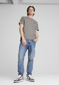 Camiseta gris de manga corta con un logo negro, combinada con jeans de mezclilla azul claros y zapatillas elegantes en colores azul marino, blanco y acentos rojos.
