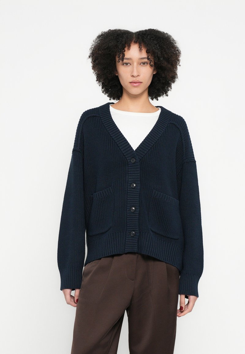 Tengerészkék kötött kardigán hosszú ujjakkal, bordázott mandzsettákkal és V-nyakkal. Két elülső zsebbel és öt gombbal záródik az elején.