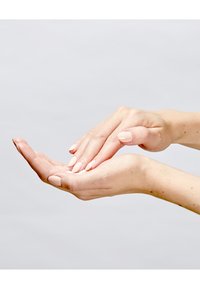 Hände mit hellroten Nägeln tragen eine Creme auf eine Hand auf und zeigen dabei glatte Haut vor einem hellgrauen Hintergrund.