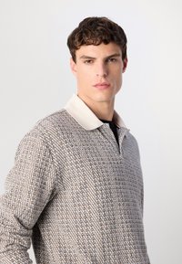 Young man with short curly hair wearing a beige and black patterned long-sleeve shirt with a white collar, looking straight ahead.