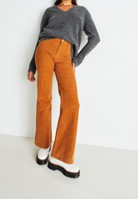 Osoba v šedém svetr s výstřihem do V, vysokých spadnutých kachníkových kalhotách v barvě spáleného pomeranče a bílých platformových botách, stojící proti bílé zdi.