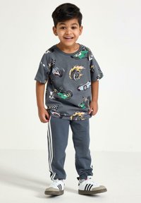 Szara koszulka T-shirt z kolorowymi wzorami samochodów i płomieniami, połączona z szarymi spodniami dresowymi z białymi paskami po bokach. Białe i szare sneakersy.