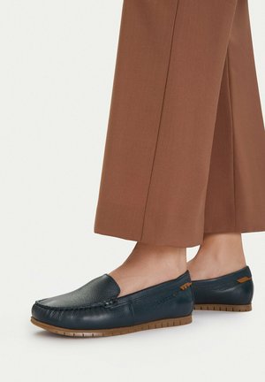 Mocasines de cuero azul marino con suelas de goma marrón, usados con pantalones marrones recortados.